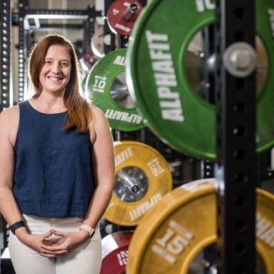 A woman stands smiling in a gymnasium, beside large colourful weights.